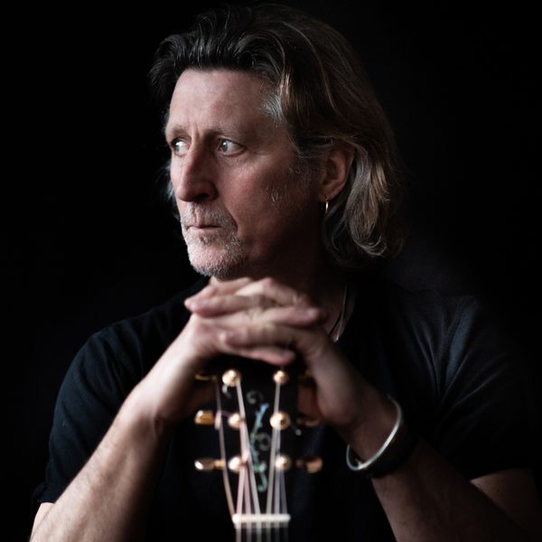 Musician Steve Knightley holding the top of a guitar and looking to the left against a black background.