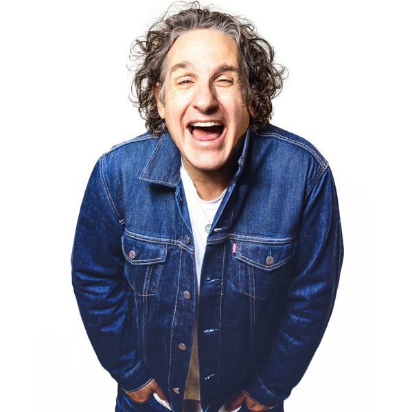 Comedian Tom Stade wearing a denim jacket leaning forwards towards the camera smiling, on a white background.