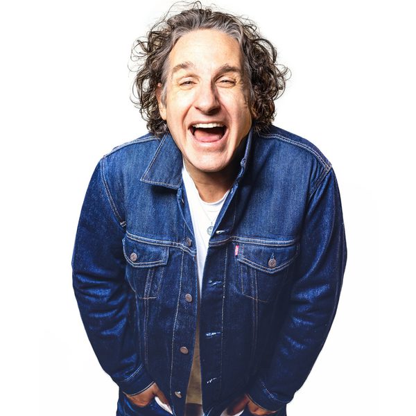 Comedian Tom Stade wearing a denim jacket leaning forwards towards the camera smiling, on a white background.