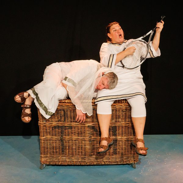 Two members of Living Spit dressed in togas and sat on a woven basket, performing onstage. One is holding a tennis racket as a banjo.