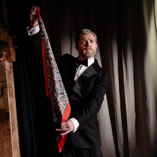 Zeb Soanes as Alec Guinness in a tux holding up a red patterned scarf against a grey theatre curtain.