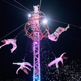 5 performers hanging off a tall structure with a dark sky, lit from below