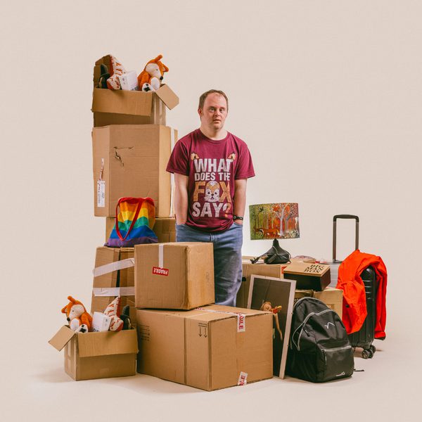 A man with Down Syndrome in a red 'What does the Fox say' t-shirt surrounded by cardboard boxes, suitcases and other belongings against a beige background.