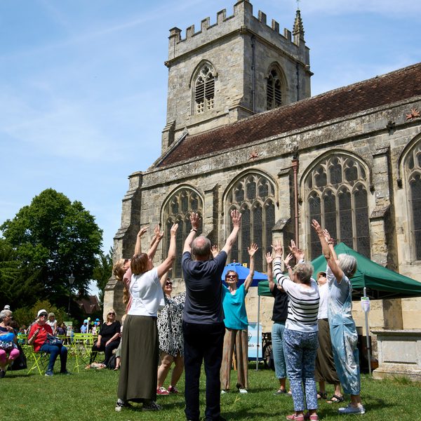 A photo of the outside of Salisbury Arts Centre with a circle of people raising their arms together in front of the building.