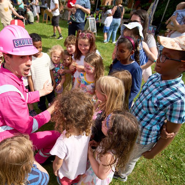 Several excited children crowded around an adult in a pink construction outfit in a green outdoor space.