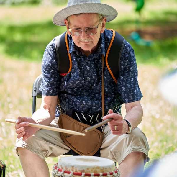 A man in a wide hat sat outside in a green space, drumming a standalone bongo.