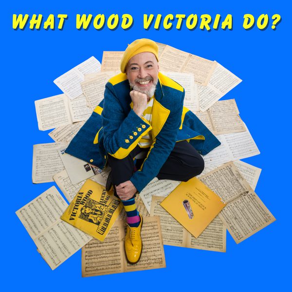 Paulus as Victoria Wood wearing a blue and yellow jacket and yellow beret sitting on a pile of sheet music. Title reads "What Wood Victoria Do?"