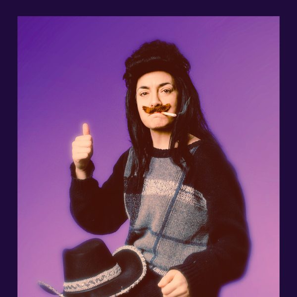 Tracey Collins dressed as Shania Wayne in a mustache and long mullet wig against a purple background, holding a cowboy hat.