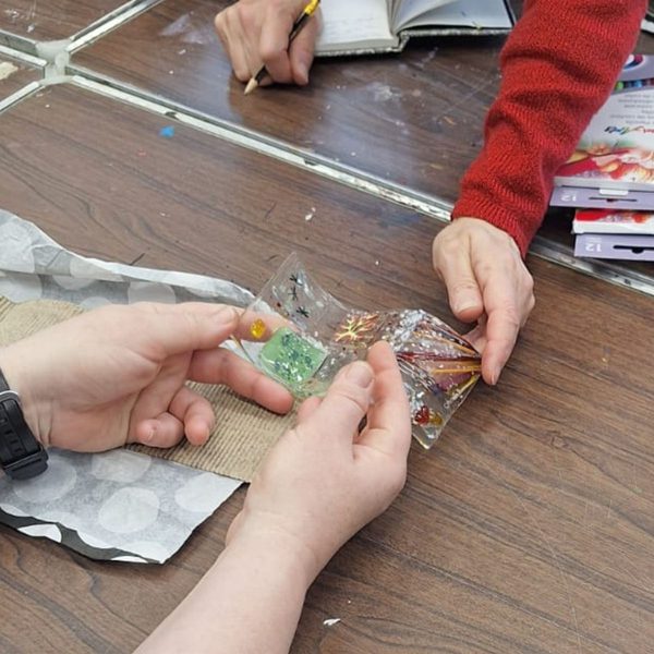 Two people reach across a table as one person hands a curved glass panel decorated with abstract patterns to the other. A notebook and art supplies are visible in the background.