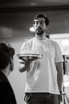 Patrick Warner standing facing a sitting Luke Barton, he is standing straight holding a tray with a decanter and glasses with a serious expression on his face.