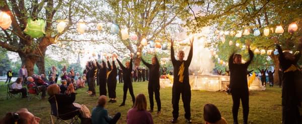 Community actors dressed in black with yellow lighting bolts on their front, stand in a line with their hands in the air. Behind them is a stage covered in lights, with festoon lanterns made from carrier bags hanging across to trees. Audience members are sat on the grass and chairs watching the performance