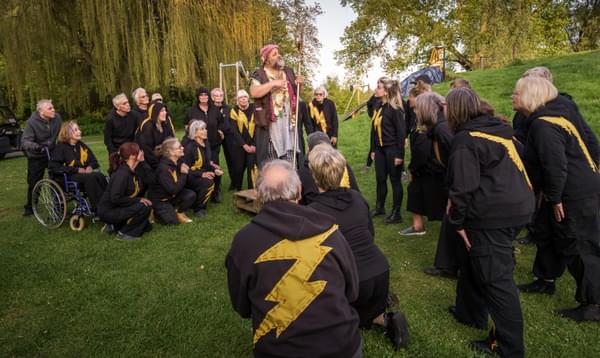 A group of actors wearing black with yellow lightning bolt crowding round Robert Bowman as Prospero, standing on a pallet