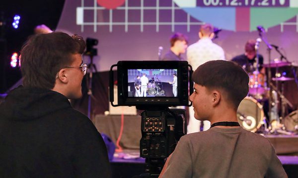 Two people operate a video camera at a live event, with a stage and a large colourful test-pattern screen in the background.