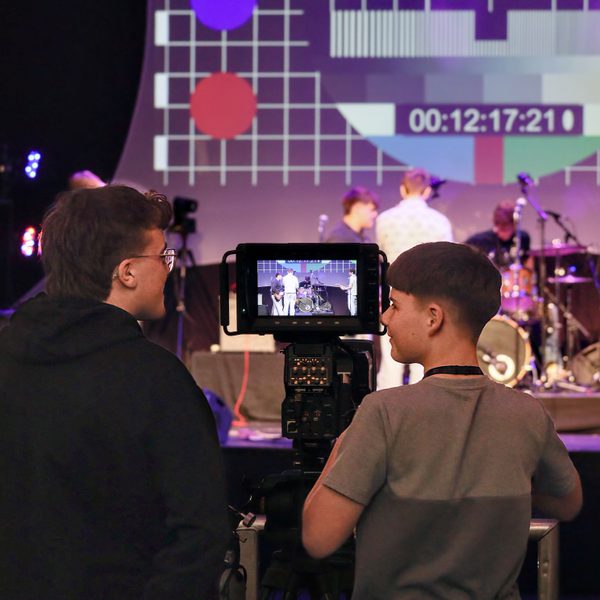 Two people operate a video camera at a live event, with a stage and a large colourful test-pattern screen in the background.