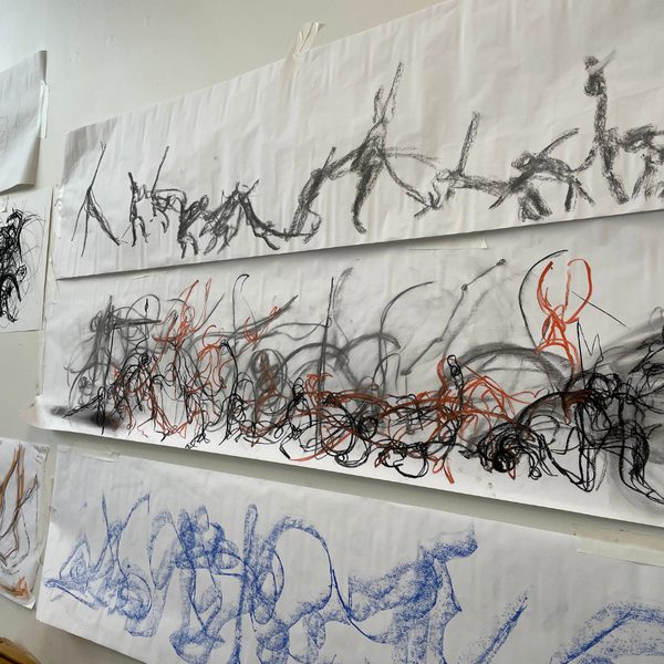 Large sheets of paper on a wall displaying abstract charcoal and pastel drawings with energetic lines, figures and sweeping marks in black, orange and blue.