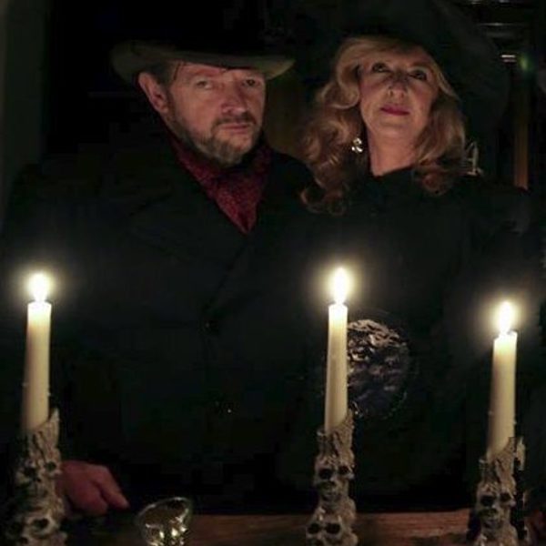Two people dressed in dark vintage-style clothing and wide-brimmed hats stand behind a wooden table lit by three tall candles in ornate skull-shaped holders.