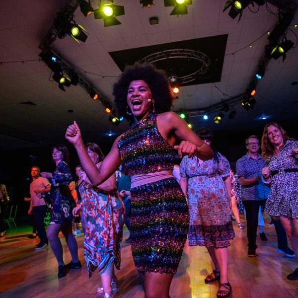 A woman in a sequinned dress leading a crowd in an energetic dance.