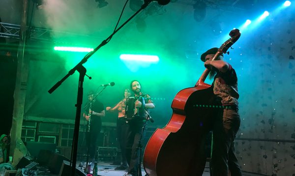 A live band performs on stage with green and blue lighting. In the foreground, a musician plays a large upright double bass. In the background, other musicians play clarinet, accordion and violin, with microphones, cables, and stage equipment visible