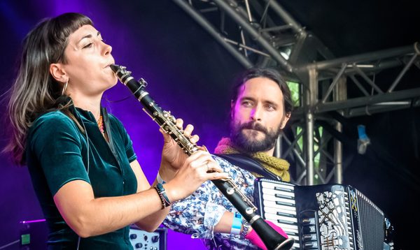 Two musicians perform on stage under purple lighting. One plays a clarinet in the foreground, while another plays an accordion slightly behind. Metal stage trussing and amplifiers are visible in the background