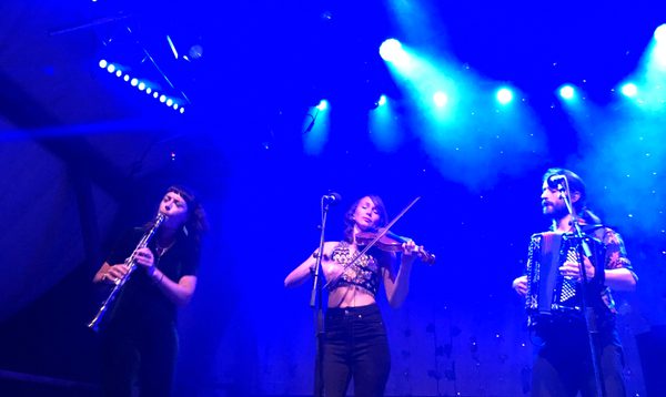Three musicians perform on a stage under intense blue lighting. One musician plays a clarinet, one plays a violin at a central microphone stand, and one plays an accordion. Stage lights and haze are visible overhead