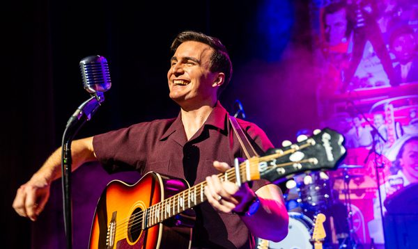 A musician playing an acoustic guitar beside a vintage microphone on a stage lit with purple and blue lights, with drums visible in the background.