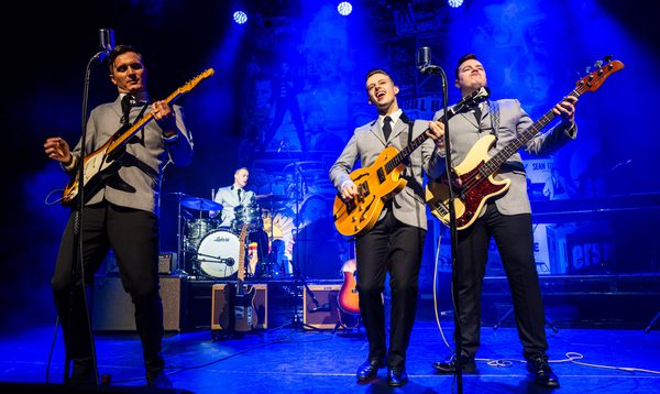 Three musicians dressed in 1960s era costumes playing guitars on stage under blue lights, with a drummer performing behind them.