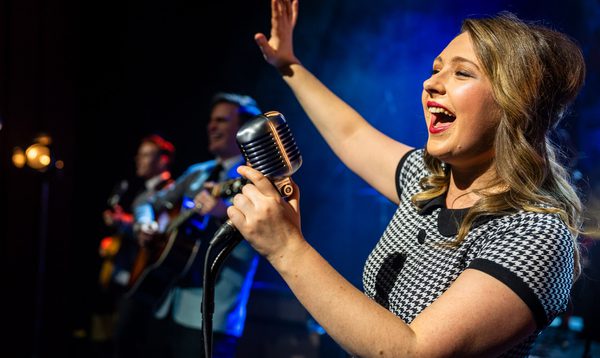 Person singing into a vintage microphone on stage, with guitarists performing in the background under blue lighting.