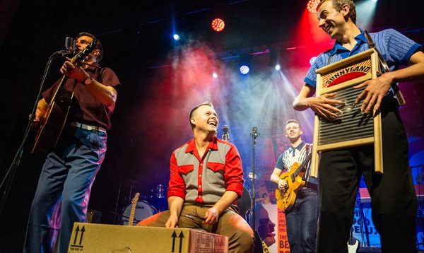 Four musicians performing on stage under colourful lights, playing guitar, percussion and a washboard.