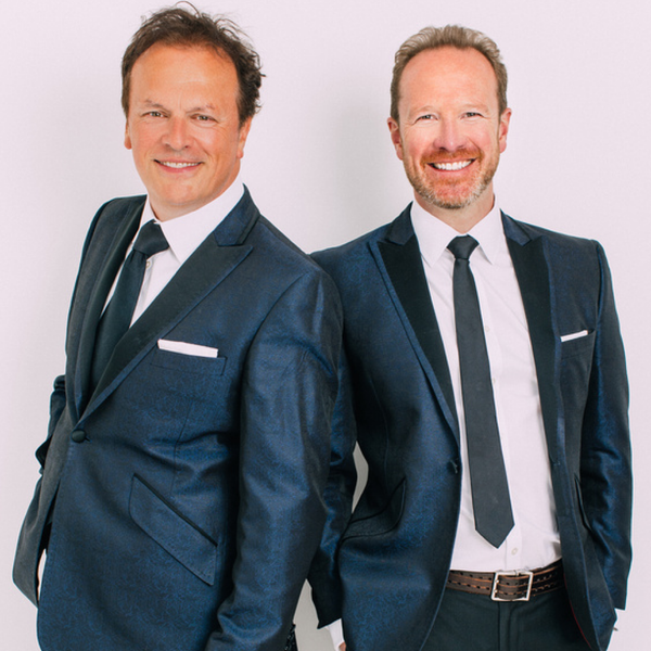 Two people standing back‑to‑back against a white studio background, wearing dark blue tailored suits with white shirts and dark ties.