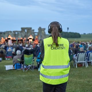 A volunteer wearing a Wiltshire Creative hi-vis jacket standing behind the audience watching a performance in front of Stonehenge