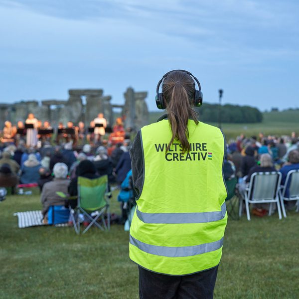 A volunteer wearing a Wiltshire Creative hi-vis jacket standing behind the audience watching a performance in front of Stonehenge