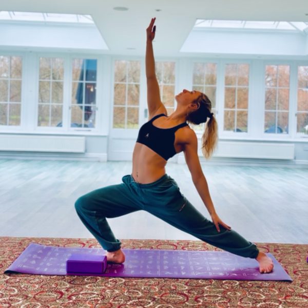 A person practising yoga on a purple mat in a bright studio, holding a wide lunge pose with one arm reaching overhead.